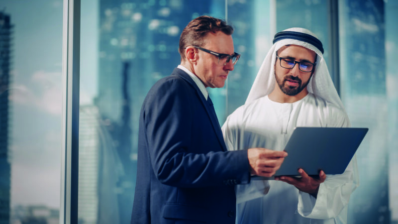 International Operations Manager Meeting Saudi Business Partner in Traditional Kandura. They’re Standing in Modern Office, Using Laptop Computer. Successful Saudi, Emirati, Arab Businessman Concept.